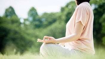 Woman sits in a grassy field with her leg crossed and hands upturned in a meditation pose 